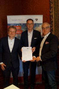 Drei Männer mittleren Alters in dunklen Anzügen stehen nebeneinander, der mittlere präsentiert eine Urkunde: