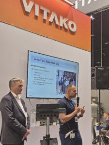 Zwei männliche Personen, einer mit Mikro in der Hand, halten einen Vortrag am Vitako-Messestand, auf einem Bildschirm sind die Vortragsfolien eingeblendet.