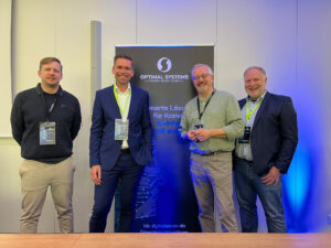 Gruppenbild mit Phillip Schulte, Björn Wittneben, Thomas Körtge und Mirko Schwellenbach vor einem Optimal-Systems-Aufsteller.