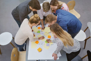 Mehrere Personen stehen über einen Tisch gebeugt zusammen und tüfteln an Post-Ist und Papierplänen.