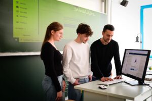 Ein Mann bedient eine Computer-Tastatur, während ein Schüler und eine Schülerin interessiert zusehen. Im Hintergrund sieht man eine digitale Schultafel.