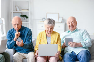 Drei ältere Personen sitzen auf einem Sofa und beschäftigen sich mit verschiedenen digitalen Endgeräten.