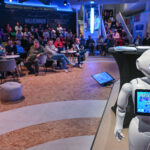 Pepper, der humanoide Roboter, steht in einem Vortragsraum, im Hintergrund sind ein Sprecher und Zuhörer zu sehen.