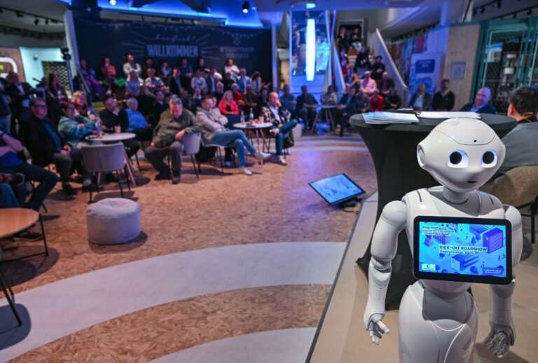 Pepper, der humanoide Roboter, steht in einem Vortragsraum, im Hintergrund sind ein Sprecher und Zuhörer zu sehen.