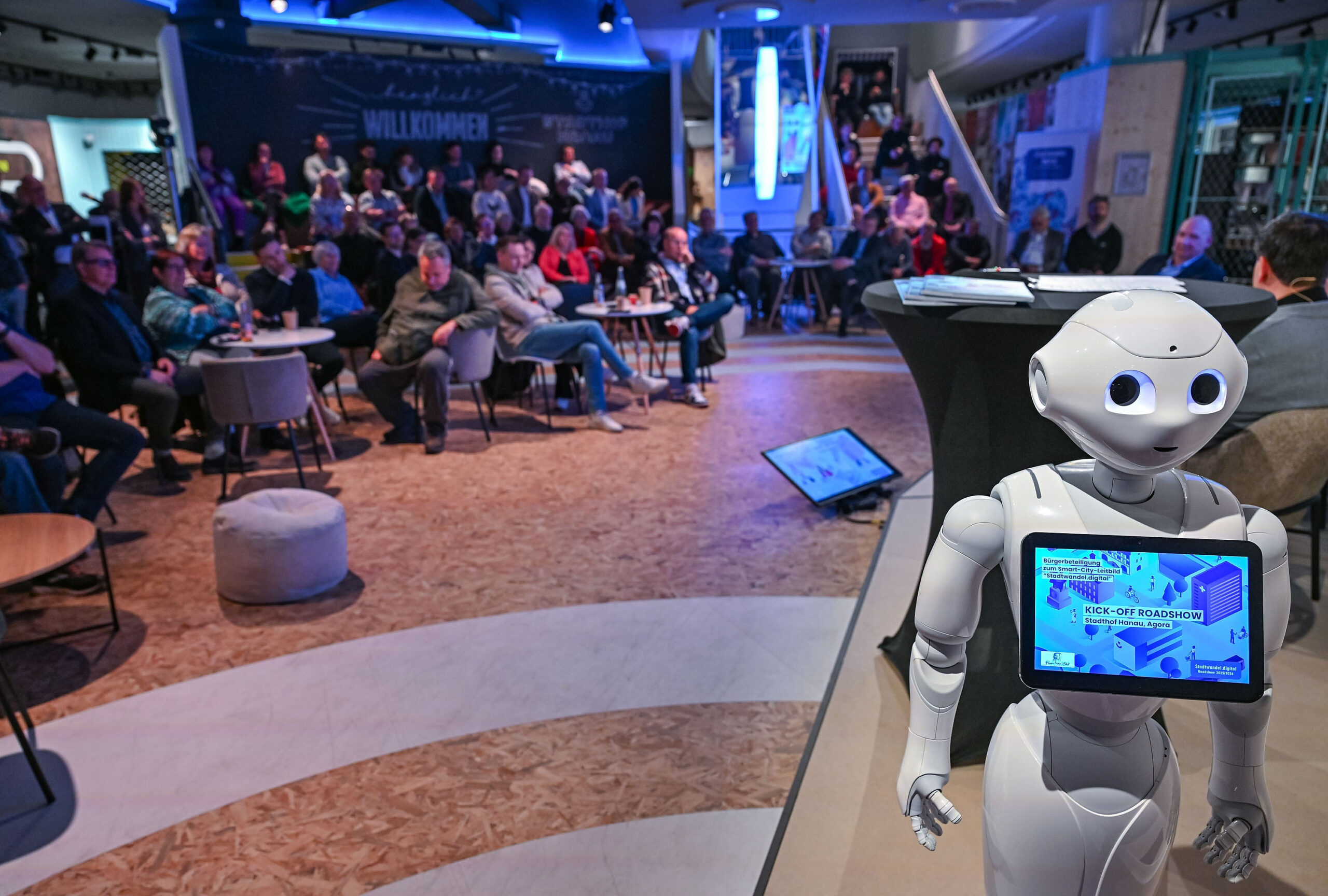 Pepper, der humanoide Roboter, steht in einem Vortragsraum, im Hintergrund sind ein Sprecher und Zuhörer zu sehen.