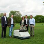 Fünf Personen stehen um den Reinigungsroboter – ein kniehohes kastenförmiges Gerät – versammelt, der auf einer Wiese im Park aufgestellt ist.