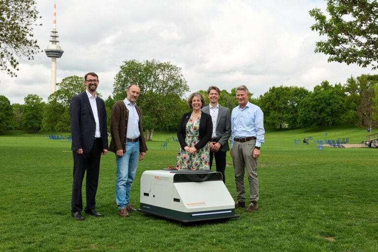 Fünf Personen stehen um den Reinigungsroboter – ein kniehohes kastenförmiges Gerät – versammelt, der auf einer Wiese im Park aufgestellt ist.