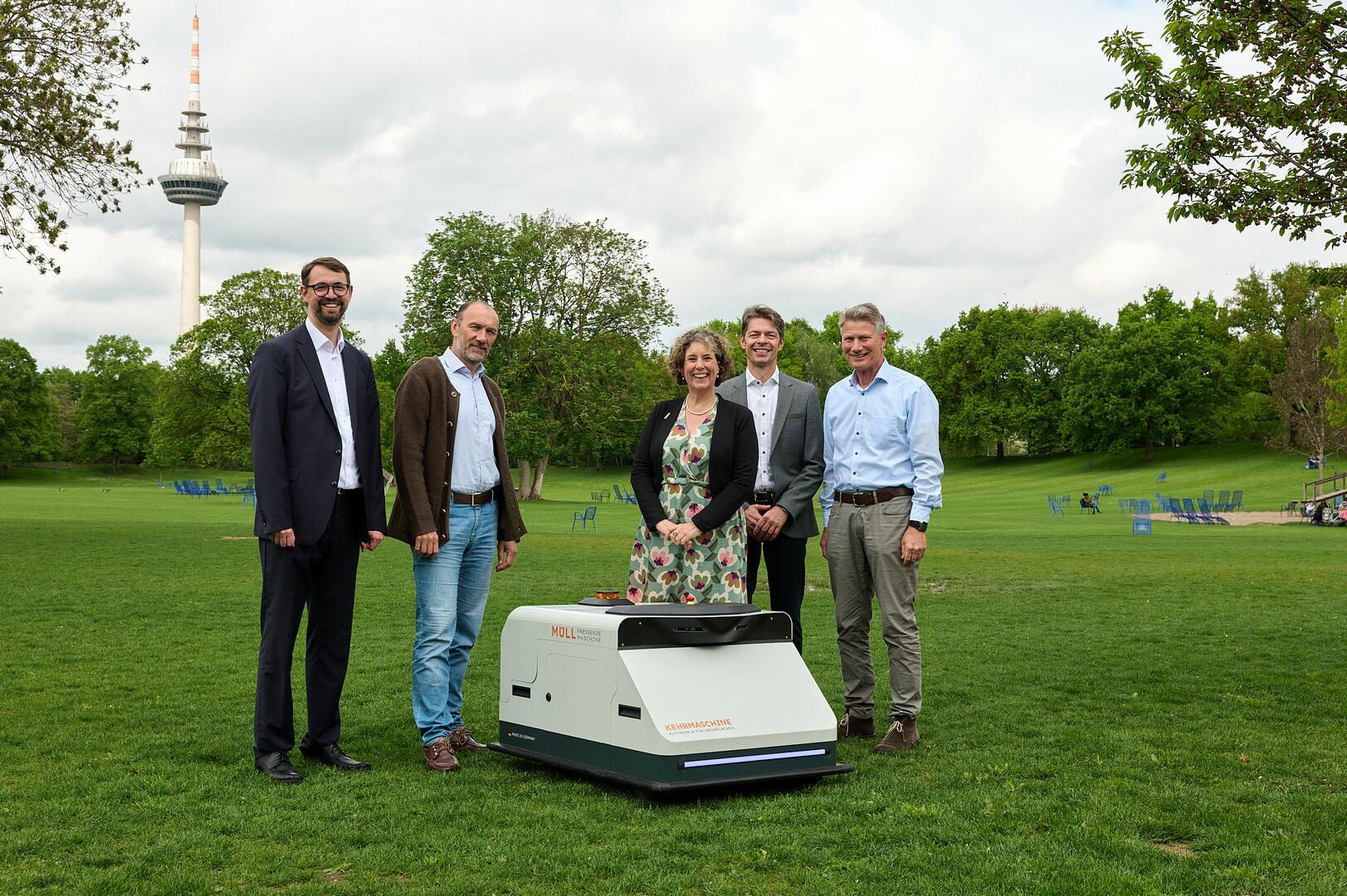 Fünf Personen stehen um den Reinigungsroboter – ein kniehohes kastenförmiges Gerät – versammelt, der auf einer Wiese im Park aufgestellt ist.