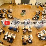 raufsicht auf einen Sitzungssaal mit runer Sitzanordnung, überlagert vom YouTube-Play-Button.