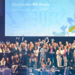Eine größere Gruppe gut gelaunter Menschen auf einer Bühne, im Hintergrund die große Projektion "Deutscher PR-Preis".
