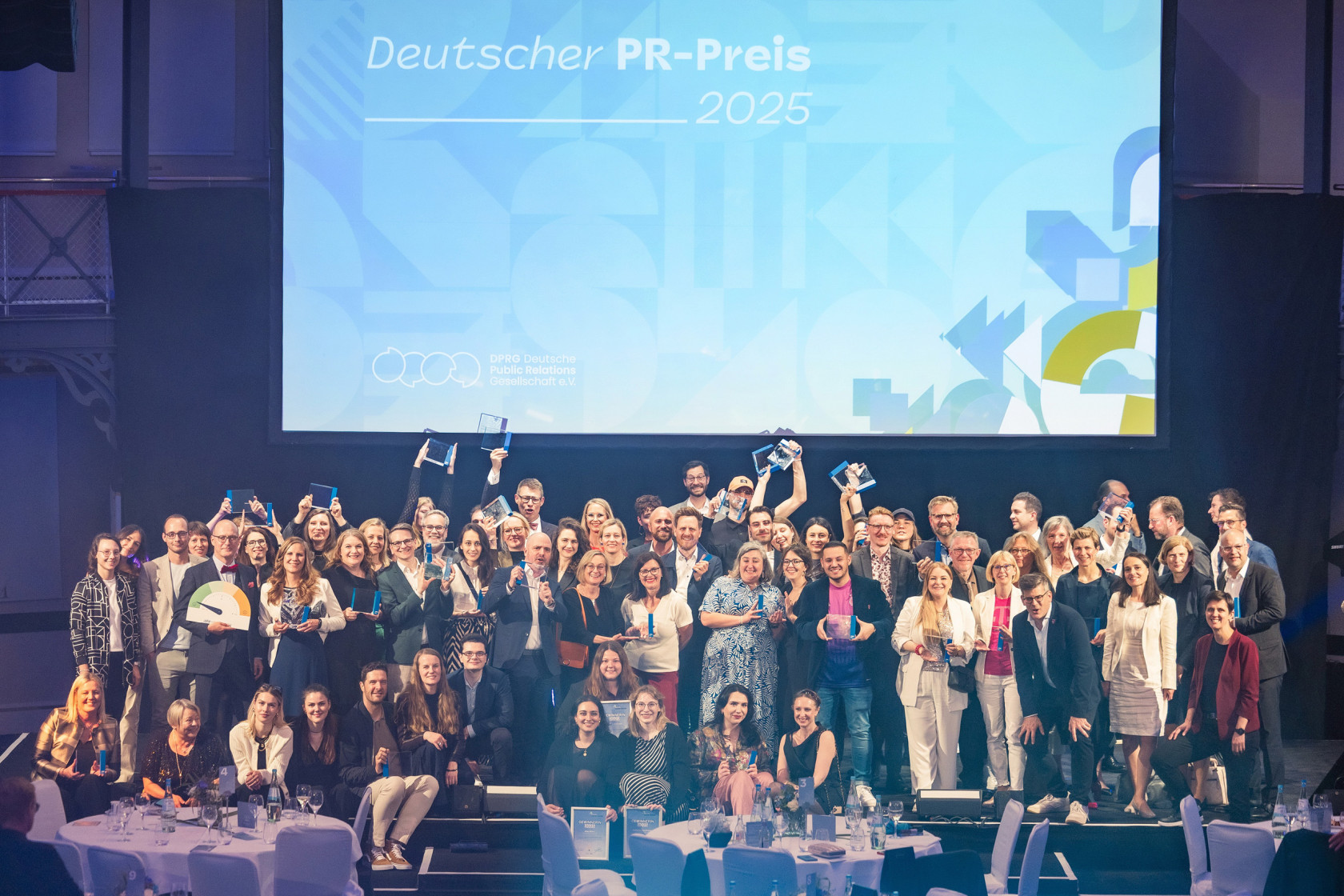 Eine größere Gruppe gut gelaunter Menschen auf einer Bühne, im Hintergrund die große Projektion "Deutscher PR-Preis".