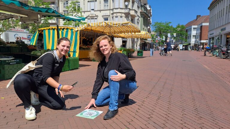 Julia Kuklik, Leiterin des Stadtarchivs Gütersloh, und Lena Jeckel, Leiterin des Fachbereichs Kultur, vor einem der QR-Codes auf dem Berliner Platz (Foto: Stadtarchiv).