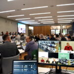 In einem großen Raum sitzen etwa 70 Personen an Tischen. Im Vordergrund ist ein großer Bildschirm zu sehen.