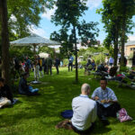 Outdoor-Szene: Konferenzbesucherinnen und -Besucher sitzen unter sommerlichem Himmel auf einer Wiese und diskutieren, im Hintergrund sind Stände zu erkennen.
