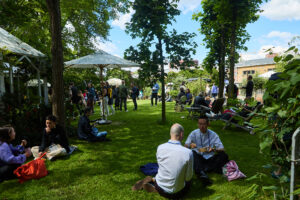 Outdoor-Szene: Konferenzbesucherinnen und -Besucher sitzen unter sommerlichem Himmel auf einer Wiese und diskutieren, im Hintergrund sind Stände zu erkennen.