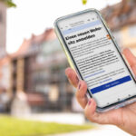 Außenaufnahme einer Hand mit Smartphone. Das Display zeigt die Website der elektronischen Wohnsitzanmeldung. Im Hintergrund ist unscharf das Erfurter Wahrzeichen, die Krämerbrücke, erkennbar.