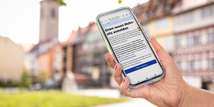 Außenaufnahme einer Hand mit Smartphone. Das Display zeigt die Website der elektronischen Wohnsitzanmeldung. Im Hintergrund ist unscharf das Erfurter Wahrzeichen, die Krämerbrücke, erkennbar.