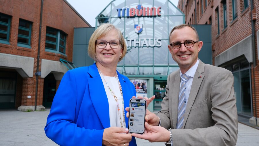 Oberbürgermeisterin Katrin Schmieder und Digitalminister Dirk Schrödter stehen nebeneinander vor dem Norderstedter Rathaus und halten gemeinsam ein Smartphone in die Kamera, auf dessen Bildschirm die neue Stadt-App zu sehen ist.