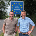 Zwei Personen stehen vor einem Straßenschild, das den Grenzübergang in die Bundesrepublik Deutschland markiert.