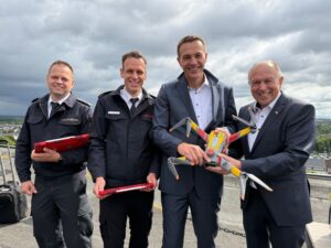 Vier männliche Personen stehen nebeneinander und halten gemeinsam eine Drohne nebst Zubehör.