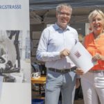 Maschine links im Bild, Mann und Frau rechts im Bild mit Papier in der Hand