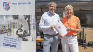 Maschine links im Bild, Mann und Frau rechts im Bild mit Papier in der Hand