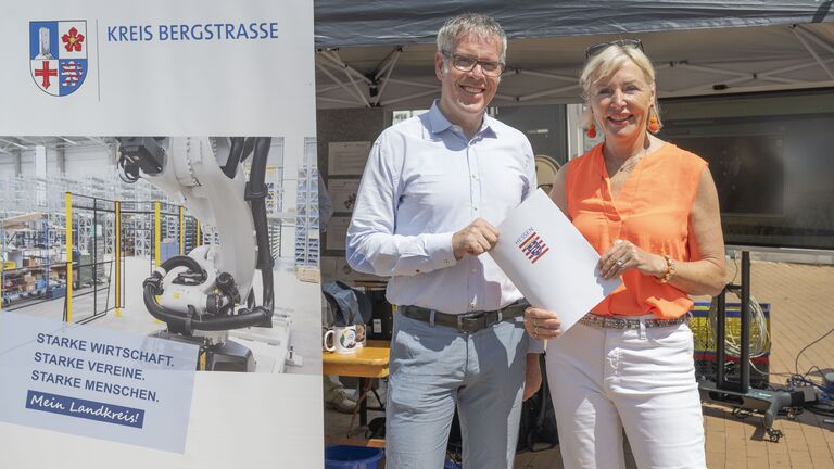 Maschine links im Bild, Mann und Frau rechts im Bild mit Papier in der Hand