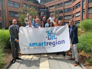 Menschen vor einem Gebäudekomplex halten das neue Logo der Smart Region Warendorf auf einem Tuchhoch