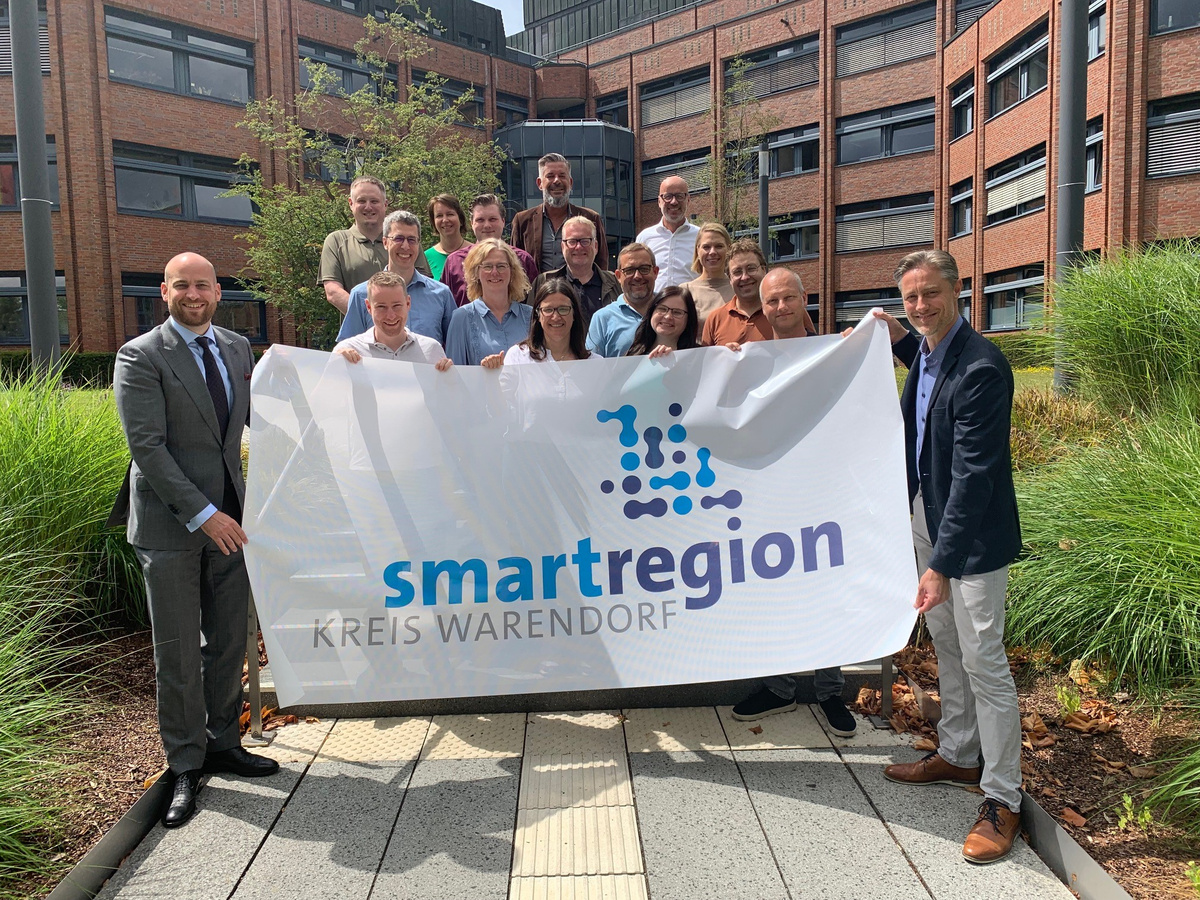 Menschen vor einem Gebäudekomplex halten das neue Logo der Smart Region Warendorf auf einem Tuchhoch
