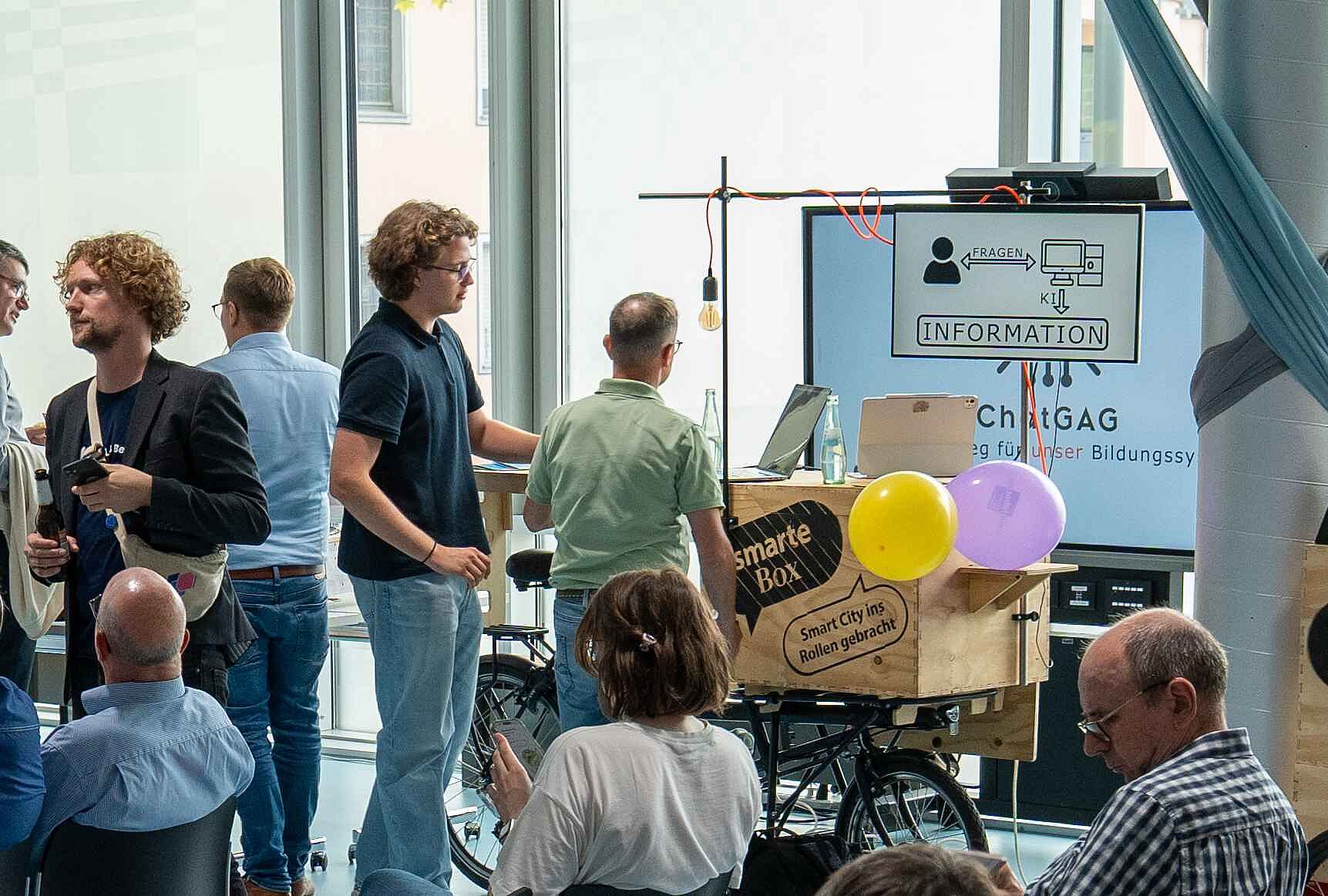 Mehrere Menschen widmen sich in einem Raum unterschiedlichen Informationstafeln und anderen Exponaten rund um die Smart City Mönchengladbach.