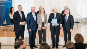 Personen auf einer Bühne, im Vordergrund sieht man Sitzreihen mit Publikum.