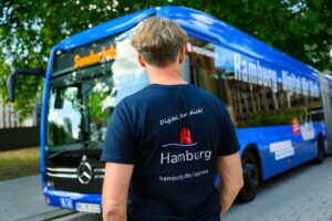 Ein Mann steht vor einem Bus. Er trägt ein Shirt mit der Aufschrift Hamburg Digital für dich, die auch auf dem Bus zu sehen ist.