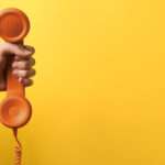 Frau hält orangefarbenen Telefonhörer vor gelbem Hintergrund