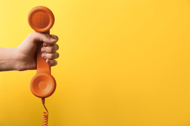 Frau hält orangefarbenen Telefonhörer vor gelbem Hintergrund
