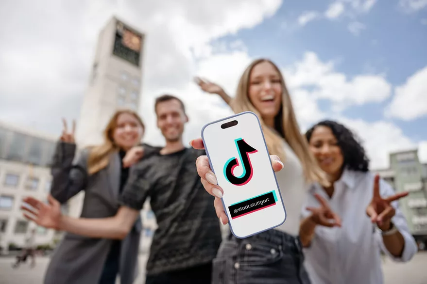 Junge Menschen stehen vor dem Stuttgarter Rathaus. In der Hand halten sie ein Smartphone mit dem TikTok Logo und dem Account-Namen @stadt.stuttgart darauf.