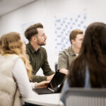 Vier Personen sitzen um einen Besprechungstisch versammelt und schauen eine Präsentation auf einem Whiteboard an.