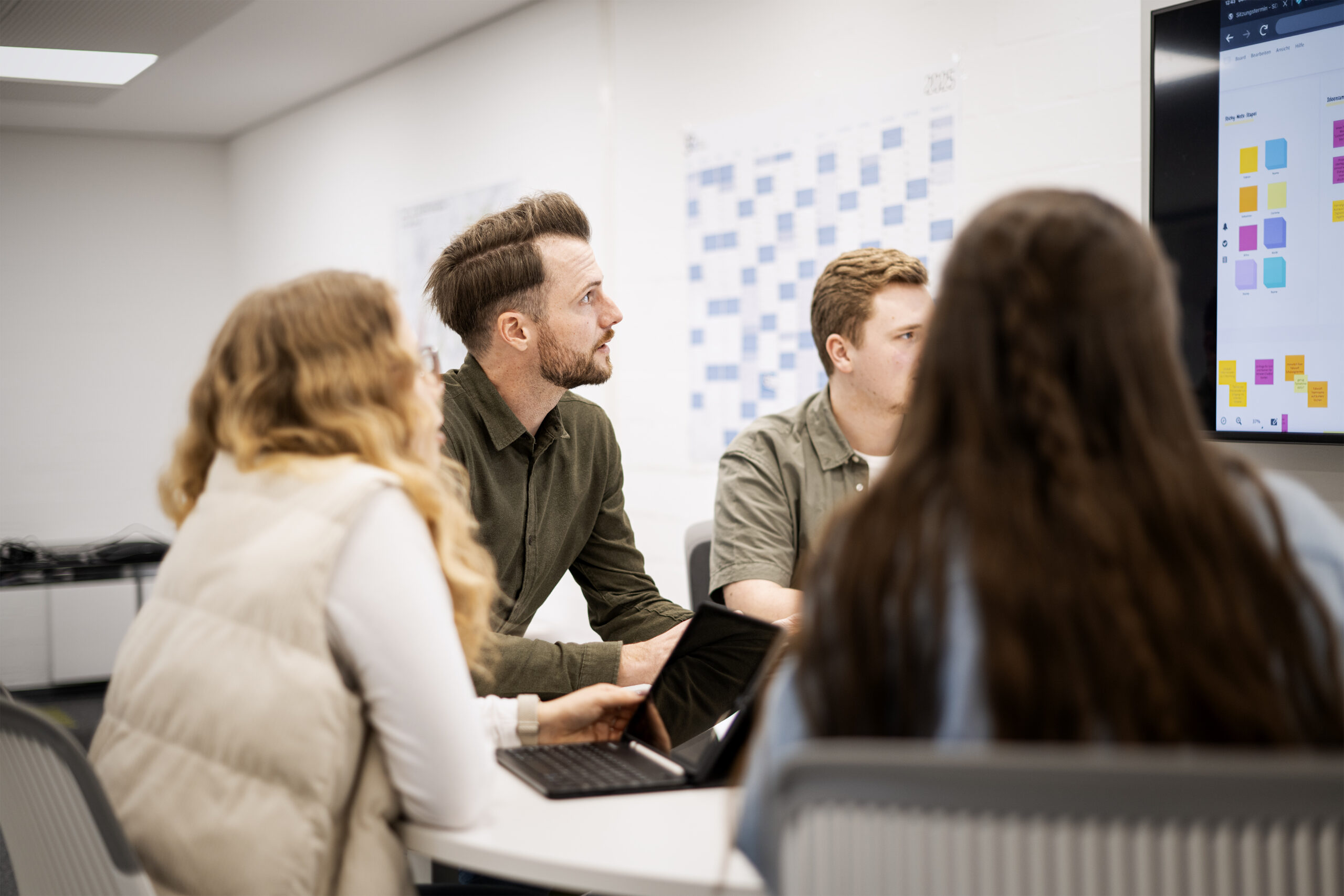 Vier Personen sitzen um einen Besprechungstisch versammelt und schauen eine Präsentation auf einem Whiteboard an.