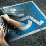 Eine Person hält mit einer Hand ein Smartphone, mit der anderen das Lenkrad eines Kfz. Im Hintergrund ist ein Behindertenparkplatz zu sehen.