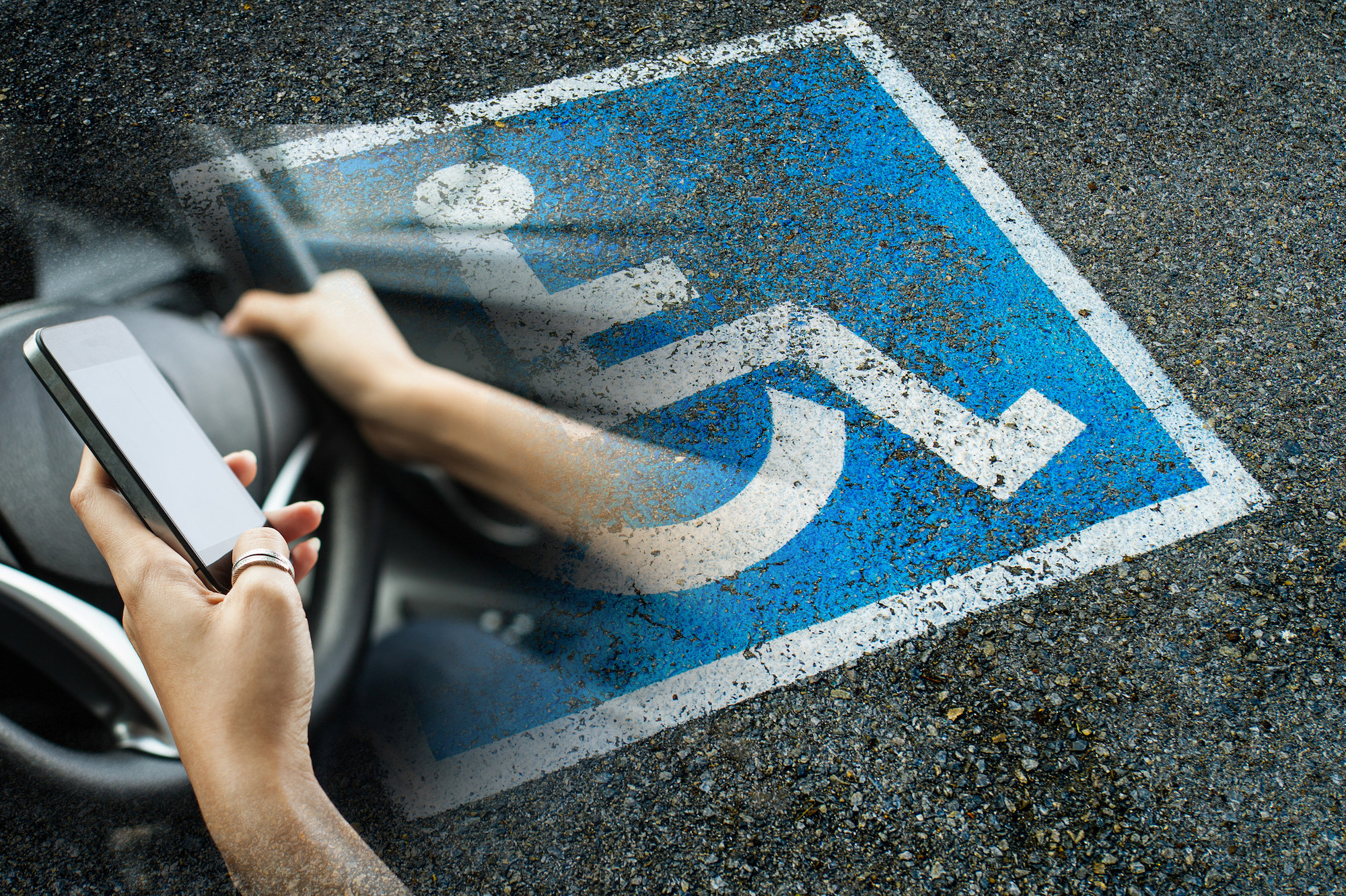 Eine Person hält mit einer Hand ein Smartphone, mit der anderen das Lenkrad eines Kfz. Im Hintergrund ist ein Behindertenparkplatz zu sehen.