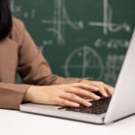 Junge Lehrerin am Laptop, Tafel im Hintergrund