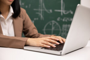 Junge Lehrerin am Laptop, Tafel im Hintergrund