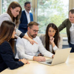 Mehrere Personen stehen um einen Tisch versammelt und schauen gemeinsam auf den Bildschirm eines Laptops.