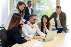 Mehrere Personen stehen um einen Tisch versammelt und schauen gemeinsam auf den Bildschirm eines Laptops.
