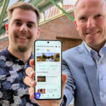 Zwei erfreut aussehende Männer stehen nebeneinander und recken ein Smartphone mit der CityApp „Gemeinde Neu Wulmstorf“. in die Kamera.