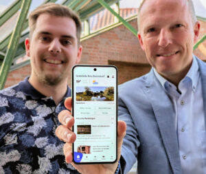 Zwei erfreut aussehende Männer stehen nebeneinander und recken ein Smartphone mit der CityApp „Gemeinde Neu Wulmstorf“. in die Kamera.