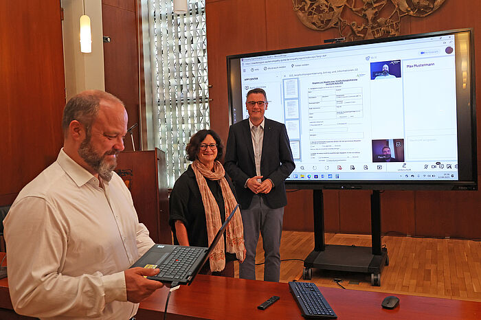 Zwei Männer und eine Frau vor einem großen beleuchteten Bildschirm zum Start des Online-Servicebüros bei der StädteRegion Aachen