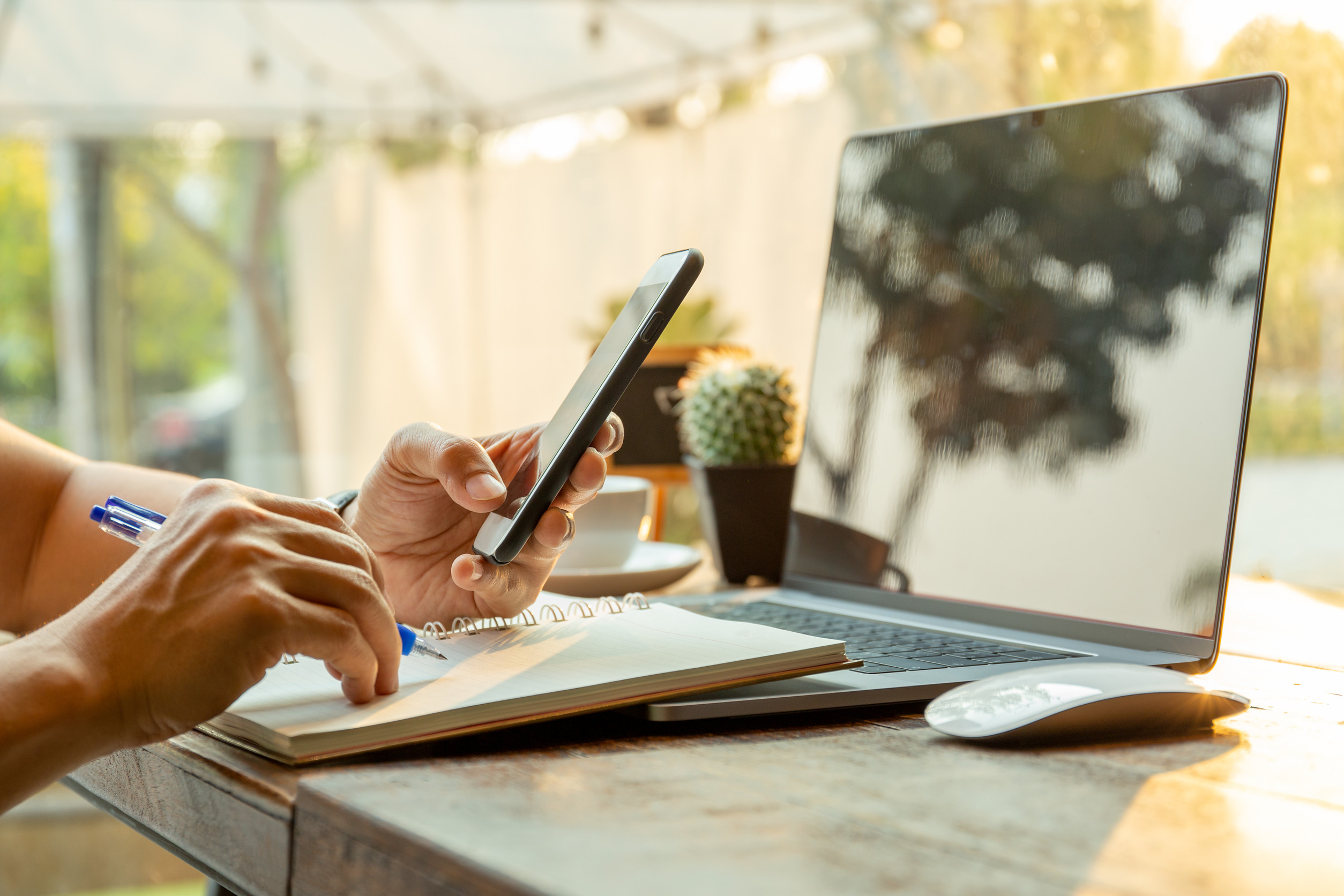 Eine Person hantiert an einem Smartphone, macht Notizen auf Papier, im Hintergrund ist ein aufgeklappter Laptop zu sehen.