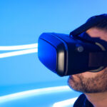 Porträtaufnahme eines jungen bärtigen Mannes mit VR-Brille vor blau-weißem Hintergrund