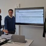 Drei Personen stehen nebeneinander vor einem Bildschirm versammelt, zwei drücken symbolisch einen Startknopf.