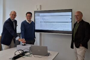 Drei Personen stehen nebeneinander vor einem Bildschirm versammelt, zwei drücken symbolisch einen Startknopf.